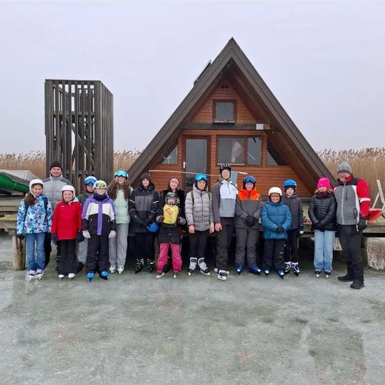 Eine Gruppe von Kindern und Erwachsenen in Winterkleidung posiert vor einer Holzhütte auf einem zugefrorenen See.