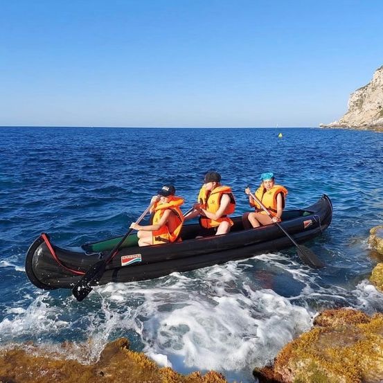 Drei Personen in orangefarbenen Rettungswesten paddeln ein schwarzes aufblasbares Kanu auf einem ruhigen Ozean mit einer felsigen Klippe im Hintergrund.