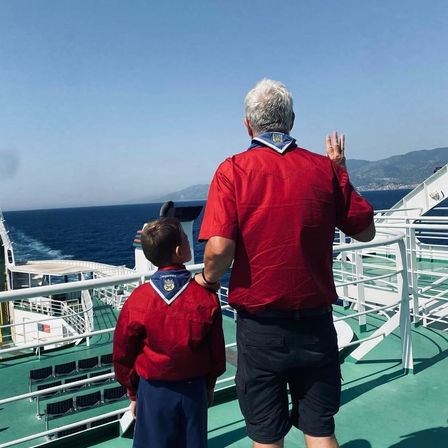 Ein Mann und ein Junge stehen auf dem Deck eines Bootes und blicken auf das Meer. Der Junge trägt ein rotes Pfadfinder-Uniform, während der Mann ein rotes Hemd und Shorts trägt.