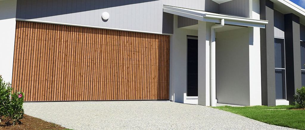Ein modernes weißes Haus mit einer hölzernen Garagentür und einer Einfahrt, die zum Eingang führt. Das Haus hat ein graues Dach, ein kleines Fenster über der Tür und eine weiße Wand.