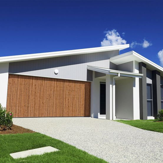 Ein modernes, helles Haus mit einer großen Holzgaragentür und einer Einfahrt, die zum Eingang führt. Das Haus hat ein schlankes Design mit weißen Wänden und einem flachen Dach.