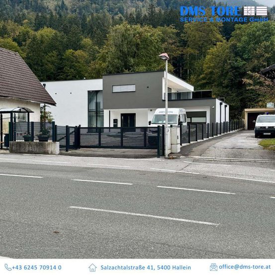 Ein Bild eines modernen Hauses mit einem schwarzen Zaun und einem weißen Van, der davor geparkt ist. Hinter dem Haus befinden sich Bäume und Berge. Unten sind Kontaktdaten und eine E-Mail-Adresse angegeben.