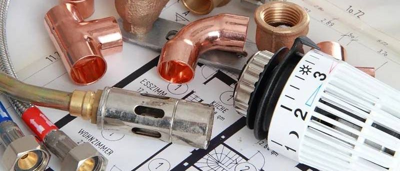 Eine Sammlung von Kupferrohren und Armaturen auf einem Bauplan. Beinhaltet einen roten Rohrschlüssel und einen Thermostat.