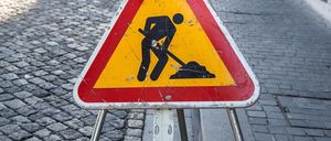 Ein Straßenschild, das auf Straßenarbeiten hinweist, steht auf dem Bürgersteig. Das Schild zeigt eine Silhouette einer Person beim Graben. Ein Auto ist in der Nähe geparkt.