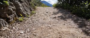 Ein Kiesweg mit Steinen und Pflanzen auf beiden Seiten, der zu einem Berg mit blauem Himmel und Wolken führt.