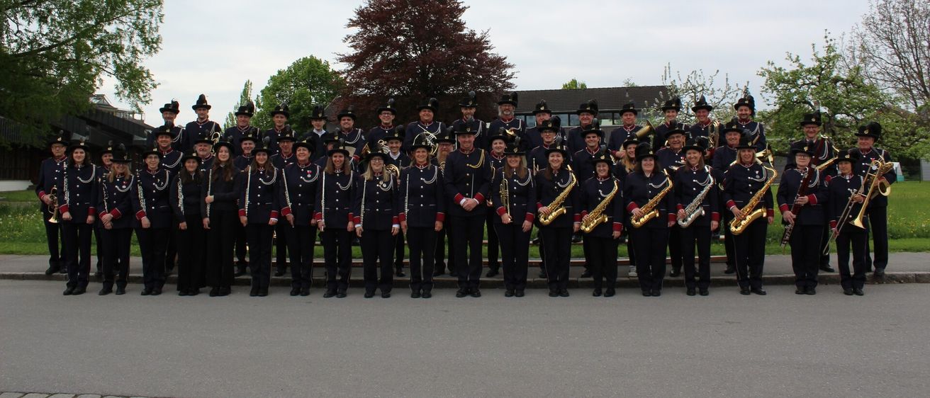 Eine große Gruppe von Musikern in Uniform posiert für ein Foto auf einer gepflasterten Fläche mit einem Haus und Bäumen im Hintergrund.