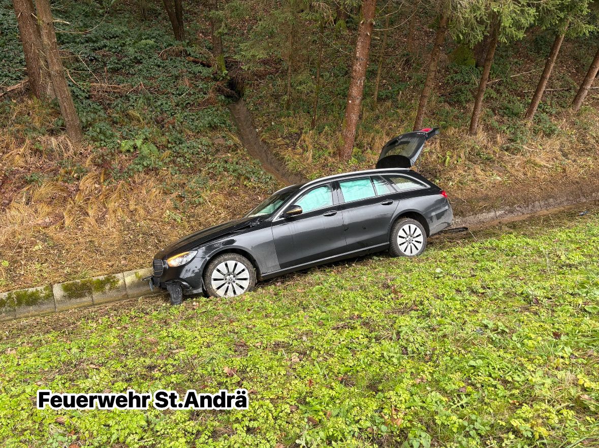 Ein schwarzer Wagen mit offenem Kofferraum steht am Straßenrand, möglicherweise nach einem Unfall. Das Auto befindet sich auf einem Grashang in der Nähe von Bäumen.
