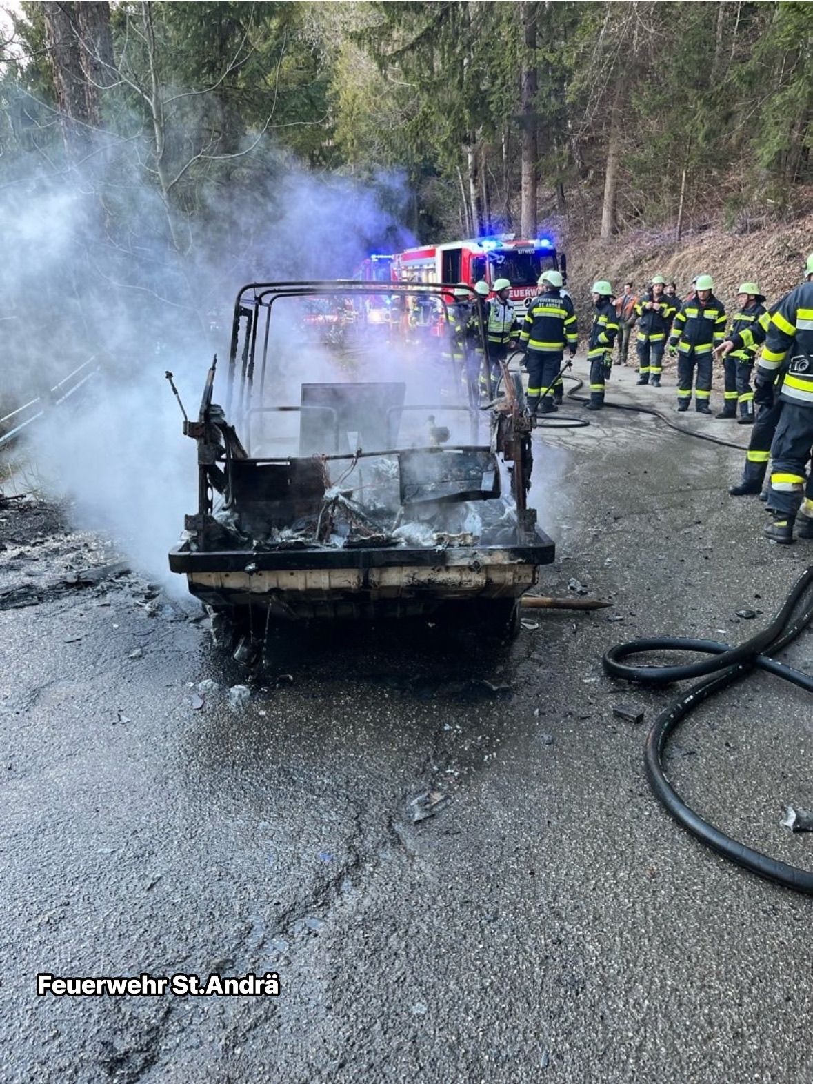 Feuerwehrleute stehen um ein verbranntes Fahrzeug auf einer Straße, aus der Rauch aufsteigt. Im Hintergrund befinden sich Bäume und ein Feuerwehrwagen.