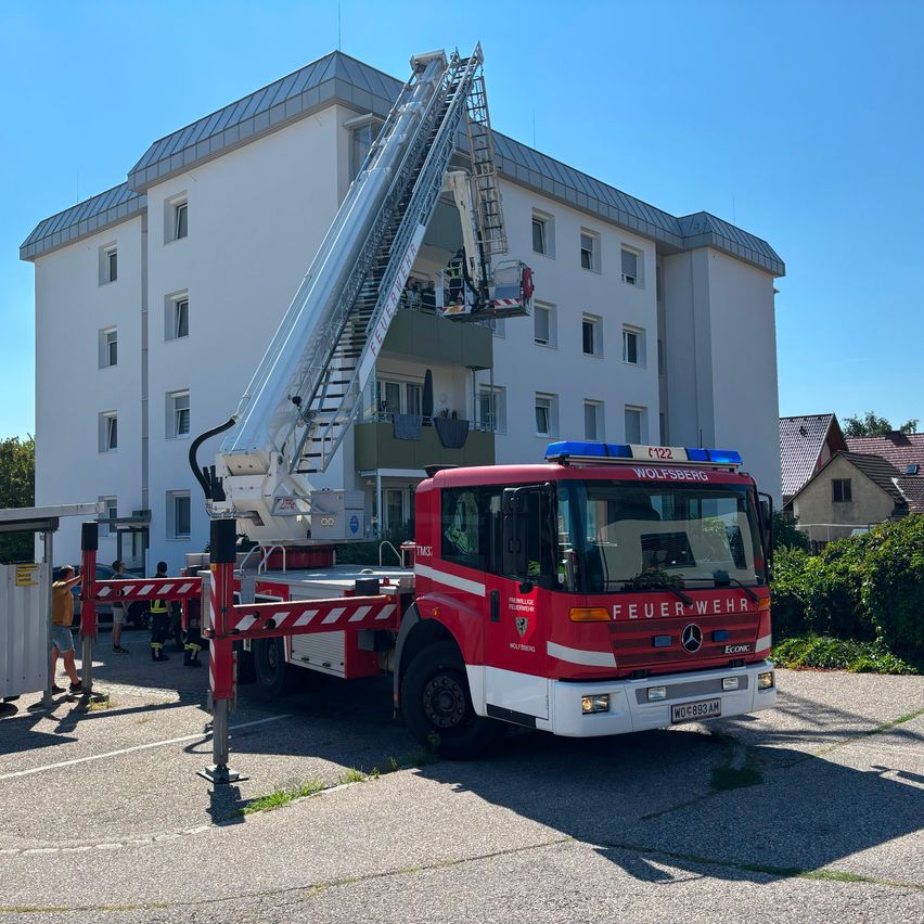 Ein roter Feuerwehrwagen mit ausgefahrener Leiter steht vor einem weißen Gebäude mit mehreren Fenstern. Eine Person steht in der Nähe des Fahrzeugs.