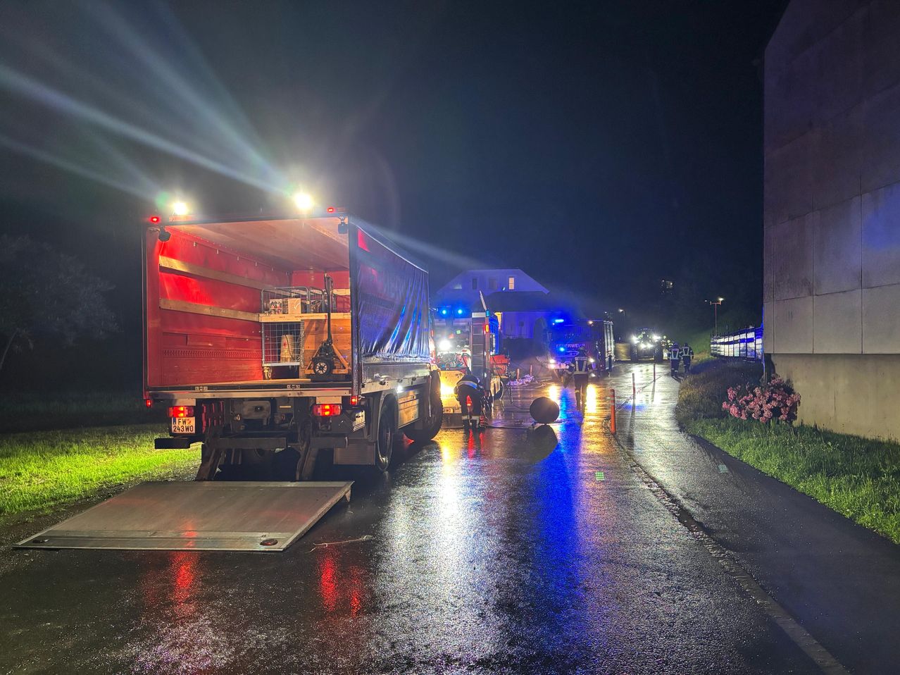 A large truck is parked on a wet road at night. It has a red back and blue lights on. A person in a helmet is fixing something near the truck. There are other vehicles and a house in the background.