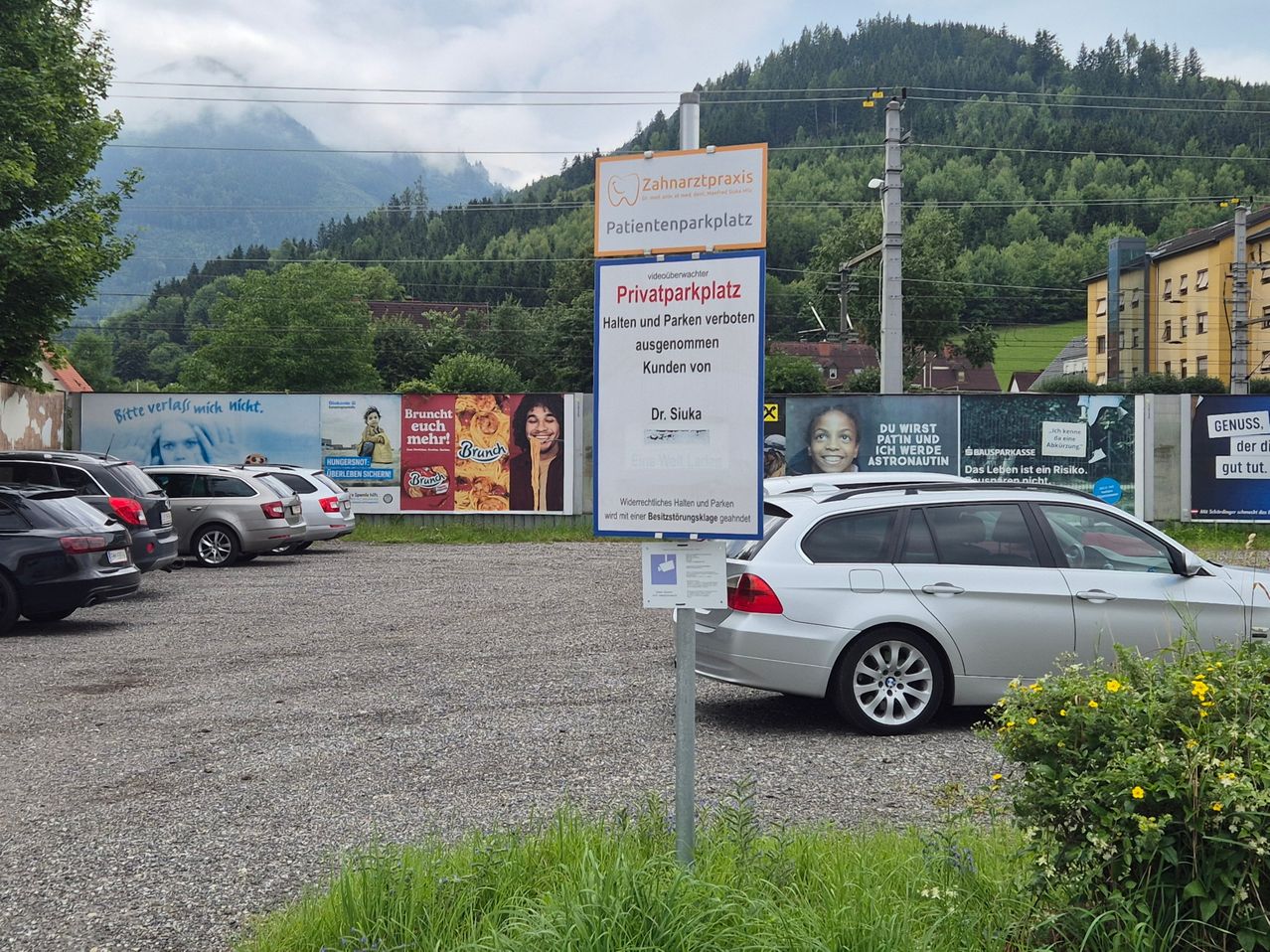 A parking lot for patients with a sign for a dental practice, surrounded by various billboards and a mountain landscape. Cars are parked in the gravel lot.