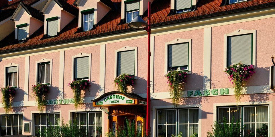 Ein rosafarbenes Gebäude mit dem Wort Gasthof am Eingang, umgeben von Pflanzen und Blumen, und einer Straßenlaterne an der Seite.