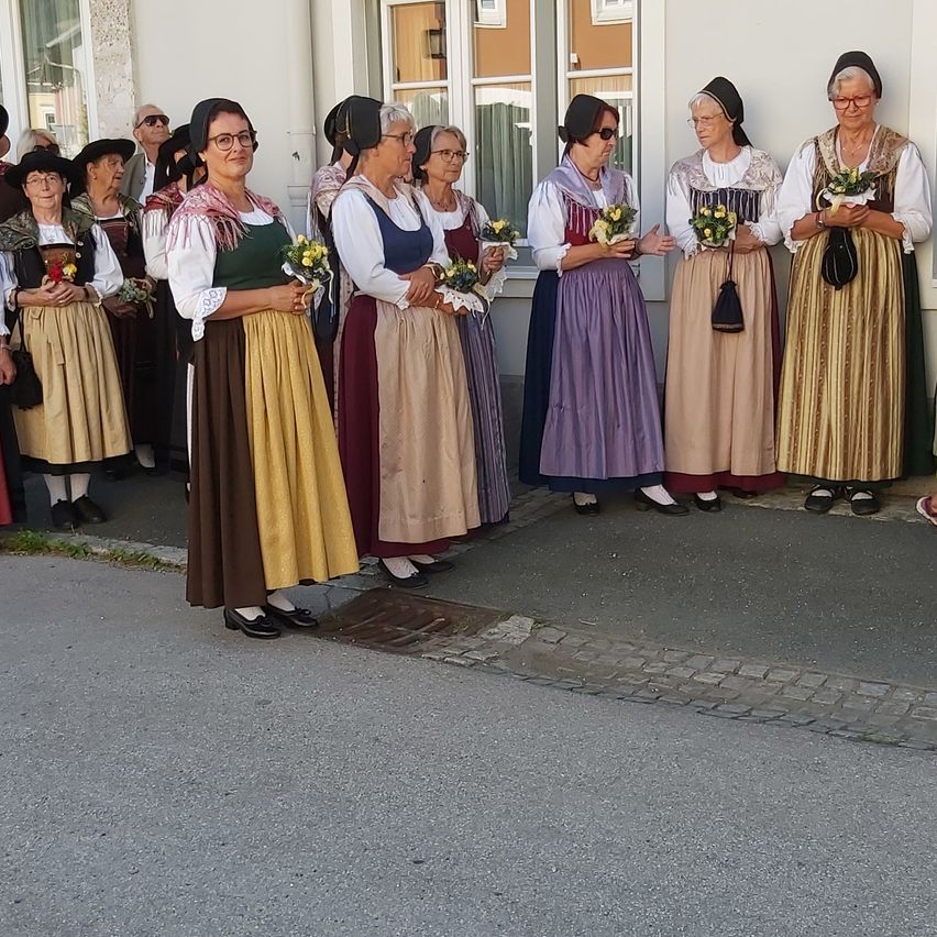 Eine Gruppe älterer Frauen in traditionellen Kostümen mit Blumen steht auf der Straße. Sie stehen vor einem Gebäude mit Fenstern.