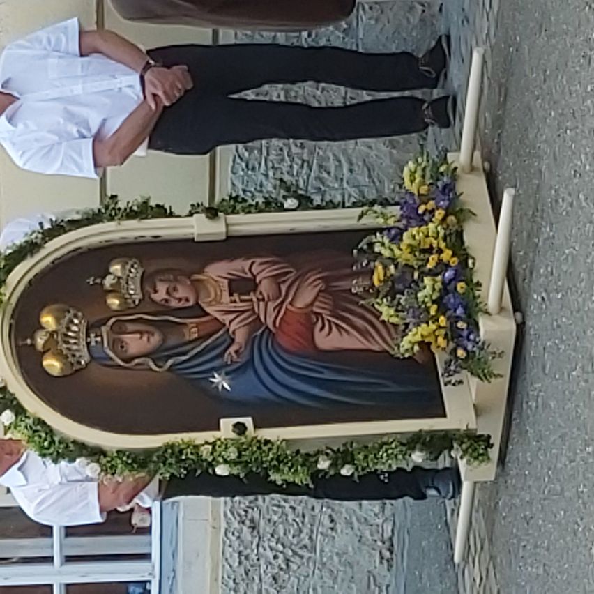 Ein religiöses Gemälde der Jungfrau Maria und Jesu steht auf dem Boden, umgeben von Blumen und Kerzen. Zwei Personen stehen in der Nähe, eine trägt ein weißes Hemd und die andere Schwarz.