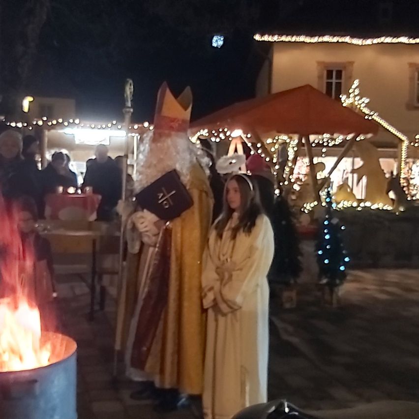 Eine Nachtszene mit einem Lagerfeuer, einem Mann als St. Nikolaus verkleidet und einem Mädchen in einem weißen Gewand. Sie befinden sich in einem festlich beleuchteten Außenbereich mit Gebäuden und einem Zelt im Hintergrund.