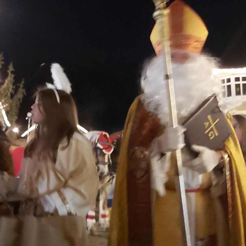 Eine als Bischof verkleidete Person führt bei Nacht eine Prozession an, hält ein Buch und einen Stab. Eine Frau in Engelsflügeln folgt ihr.