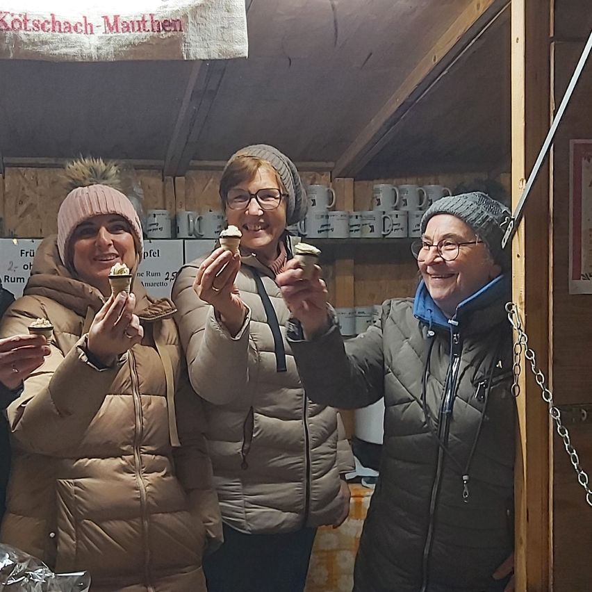 Vier Frauen halten Cupcakes in der Hand, lächeln und posieren für ein Foto in einem Geschäft. Das Geschäft hat Holzregale mit weißen Tassen. Ein Schild mit dem Wort 'Fru' ist an der Wand angebracht.