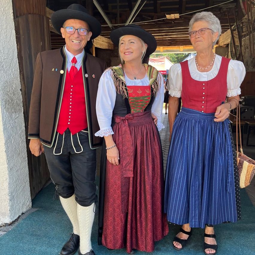 Drei Menschen in traditioneller Kleidung, zwei Frauen und ein Mann, stehen zusammen in einer rustikalen Umgebung.