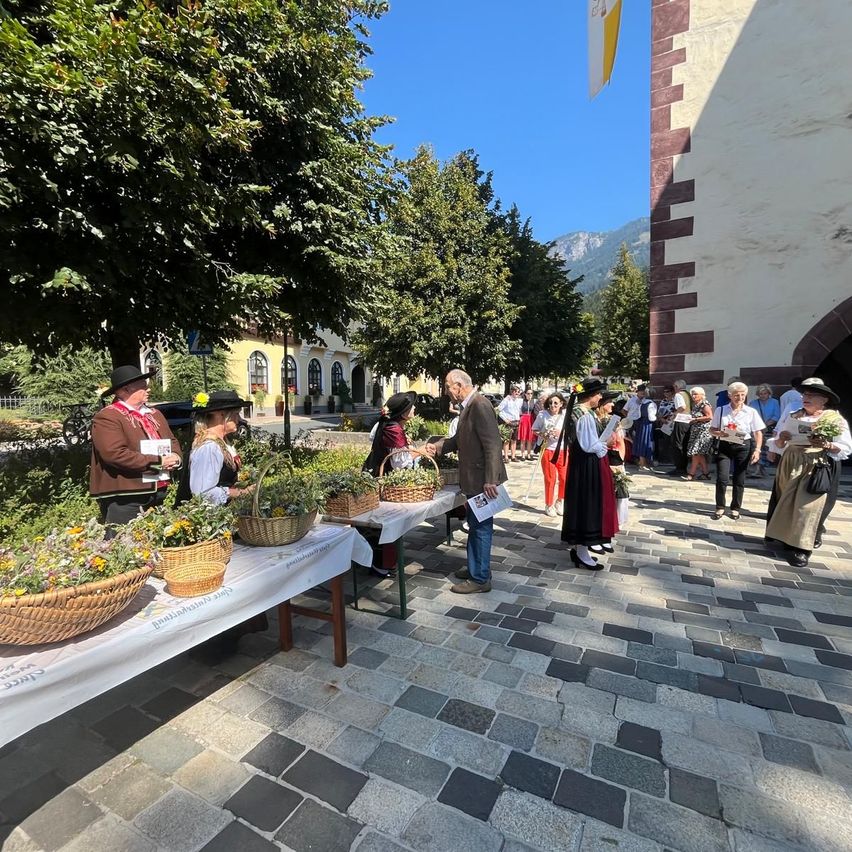 Eine Gruppe von Menschen in traditioneller Kleidung versammelt sich um einen Tisch mit Blumenkörben auf einem Stadtplatz.