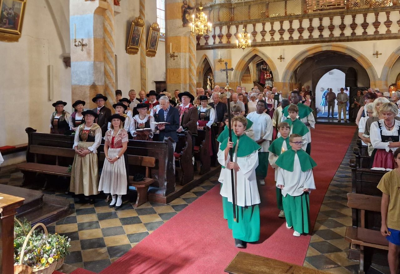 Eine Gruppe von Menschen in traditioneller Kleidung versammelt sich in einer Kirche, einige halten Bücher und andere stehen vorne mit grünen Umhängen.