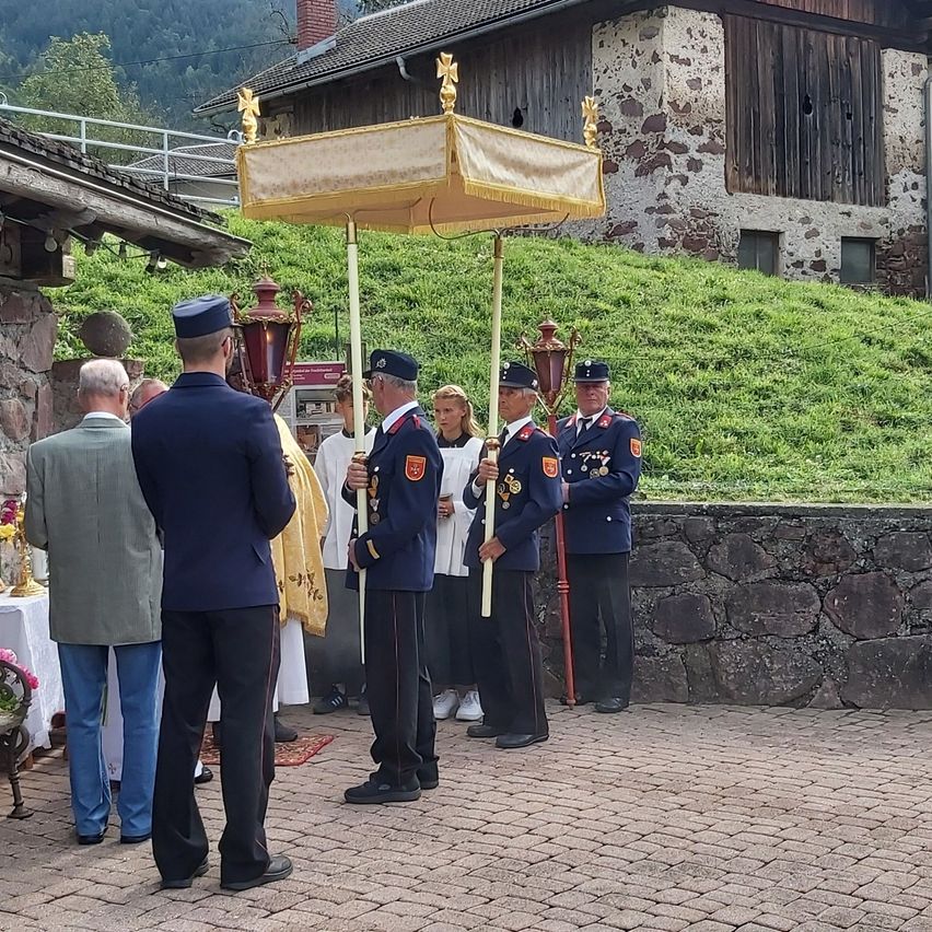 Eine Gruppe uniformierter Personen, möglicherweise Militär, steht vor einem Gebäude mit einem grasbewachsenen Hügel im Hintergrund. Sie halten zeremonielle Stäbe und nehmen an einer formellen Veranstaltung teil.