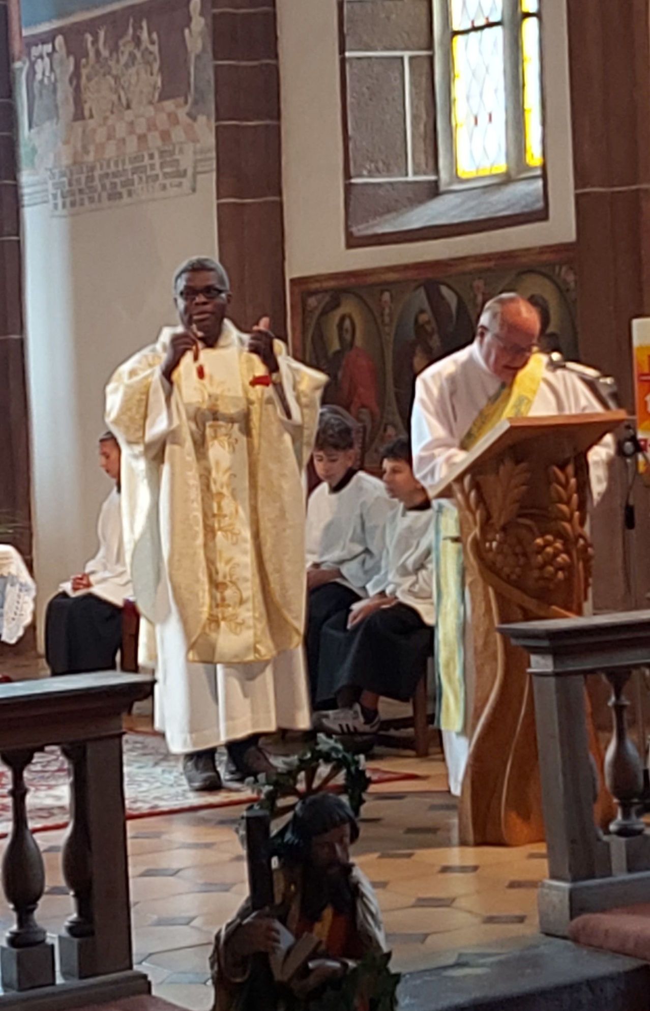 Zwei Priester stehen in einer Kirche, einer spricht am Podium, während der andere mit den Händen gestikuliert. Hinter ihnen sitzen mehrere Personen auf einer Bank.