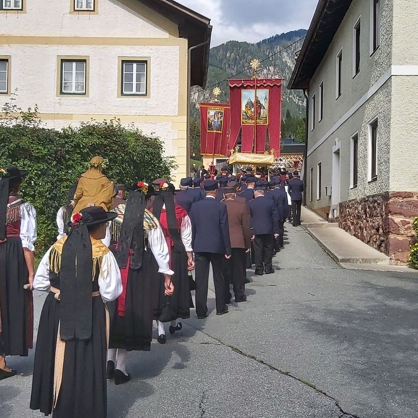 Eine Gruppe von Menschen in traditioneller Kleidung geht eine Straße entlang und folgt einer religiösen Prozession mit Bannern und einem goldenen Kreuz. Gebäude säumen die Straße, und Berge sind im Hintergrund sichtbar.
