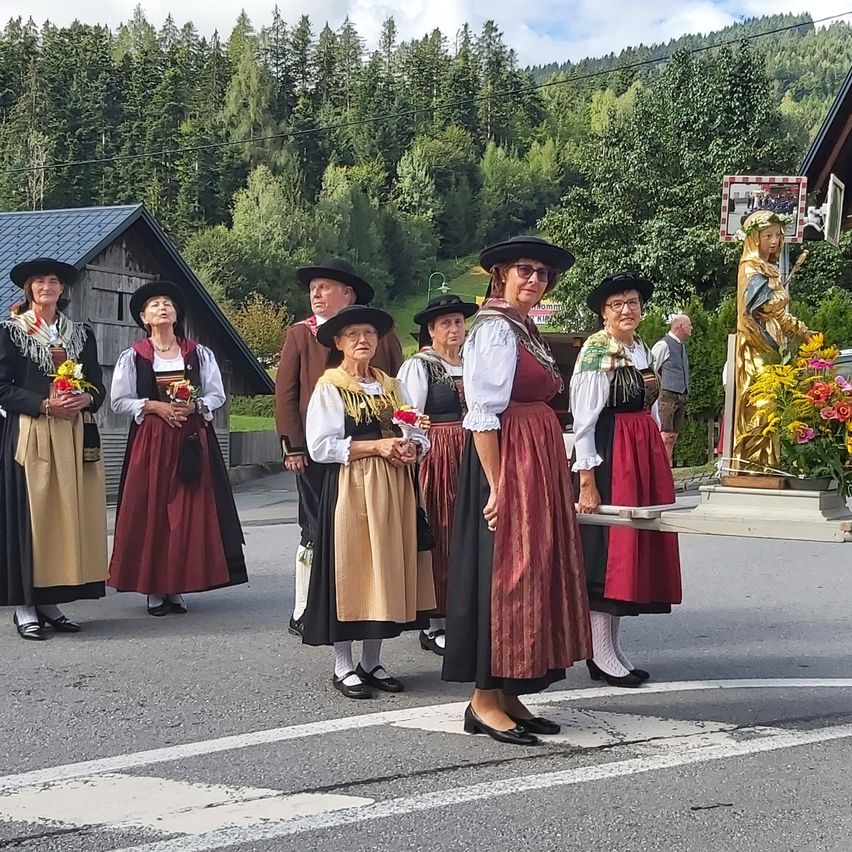 Eine Gruppe von Frauen in traditioneller Kleidung steht auf einer Straße, hält Blumen und hat eine Statue und Grünpflanzen im Hintergrund.