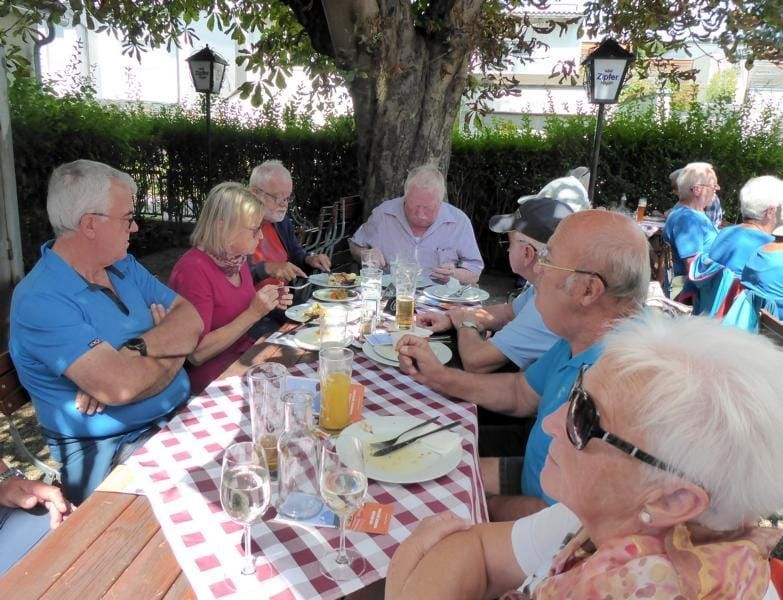 Eine Gruppe älterer Erwachsener sitzt draußen an einem Tisch. Der Tisch ist mit einer rot-weiß karierten Tischdecke bedeckt. Sie essen, mit Tellern, Gläsern und Besteck. Einige tragen eine Brille und einer eine Sonnenbrille. Hinter ihnen steht ein Baum und ein Zaun.