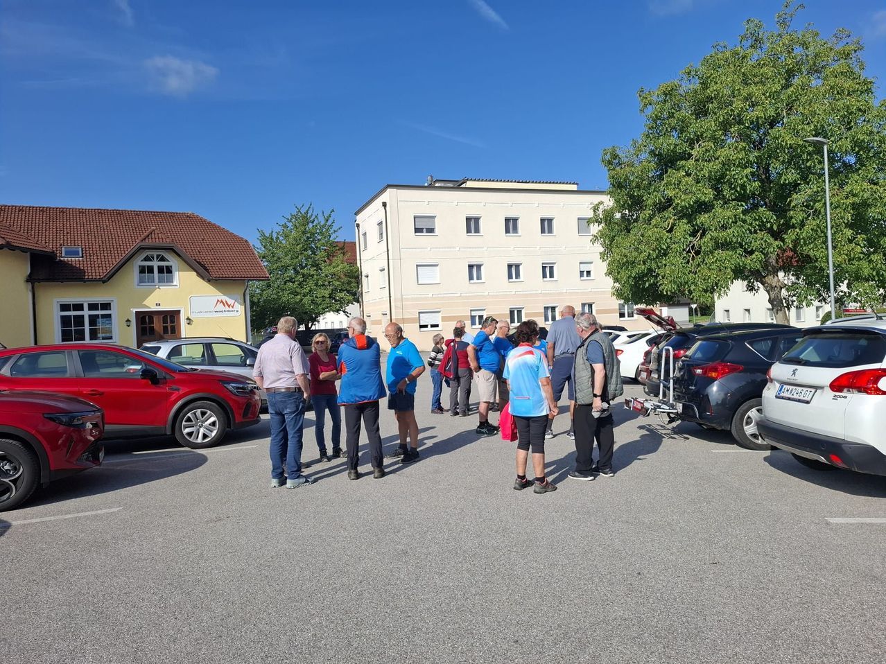 Menschen versammeln sich auf einem Parkplatz, einige tragen blaue Shirts und Sneakers. Ein Auto mit offenem Kofferraum steht in der Nähe, und ein Gebäude mit vielen Fenstern befindet sich im Hintergrund.