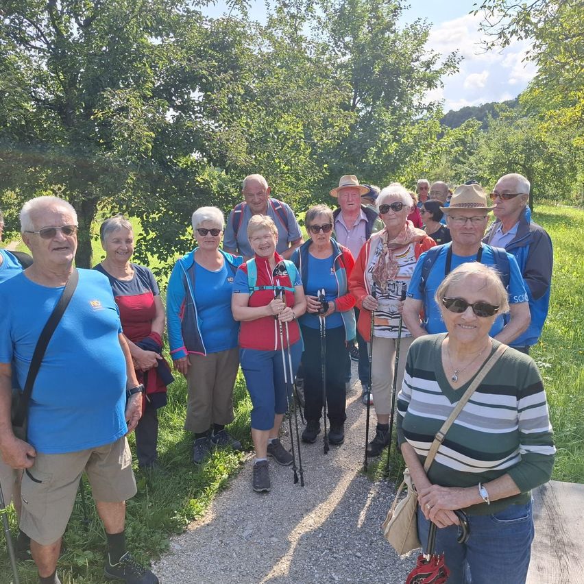 Eine Gruppe von Menschen steht auf einem Kiesweg in einem Grasbereich und trägt Sonnenbrillen und Rucksäcke. Sie alle lächeln und scheinen für ein Foto zu posieren. Der Hintergrund ist ein Wald mit Bäumen und Bergen.