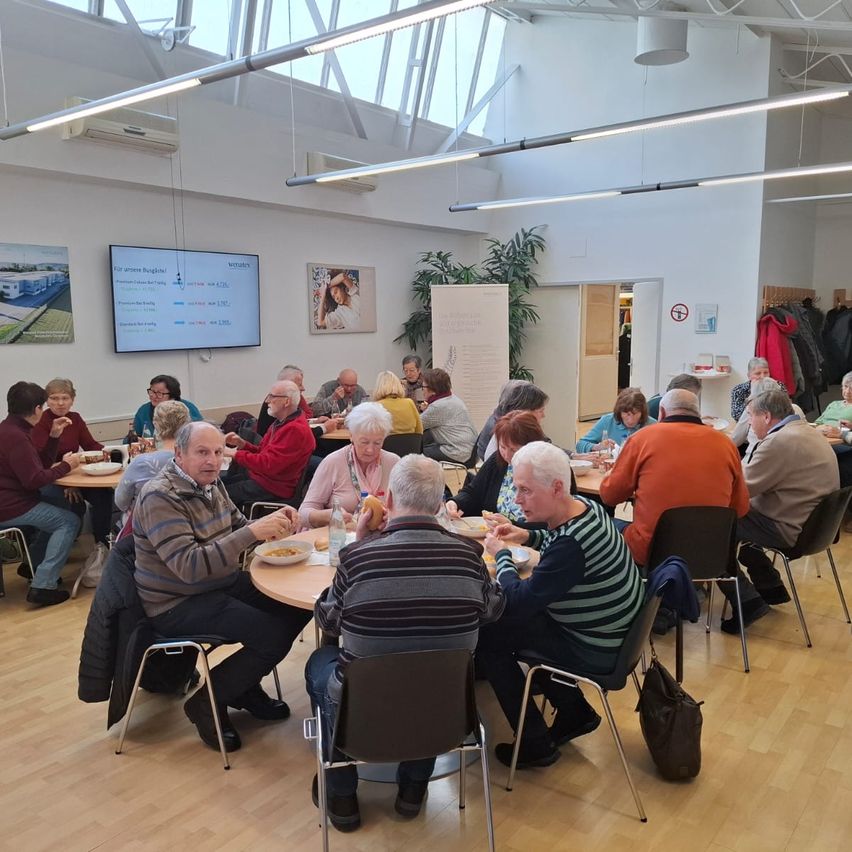 Mehrere ältere Erwachsene sitzen um Tische in einem gut beleuchteten Raum. Sie essen und plaudern. Der Raum hat Holzböden, weiße Wände und einen großen Bildschirm an der Wand.