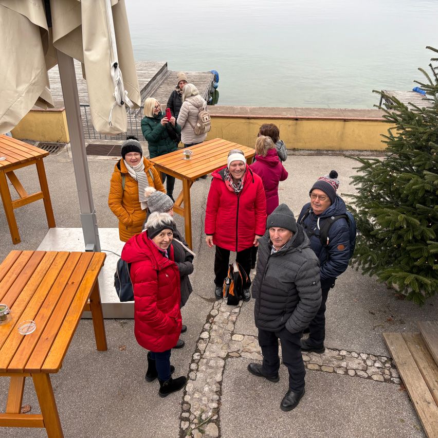 Mehrere Personen in Winterkleidung stehen bei Tischen in der Nähe eines Sees. Rechts steht ein Weihnachtsbaum.