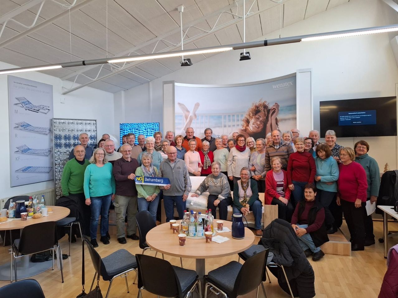 Eine Gruppe von Menschen versammelt sich in einem Raum, einige sitzen um einen Tisch mit Erfrischungen, während andere auf einem Sofa stehen oder sitzen. Ein großes Poster mit einer Frau und einem Kind ist an der Wand angebracht.