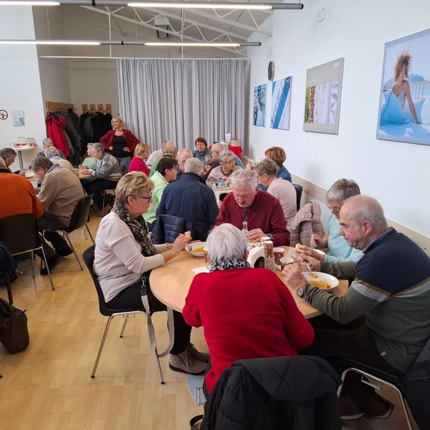 Eine Gruppe älterer Menschen sitzt um einen Esstisch in einem Raum herum und isst. Der Raum hat einen Holzboden, weiße Wände und montierte Rahmen. Es gibt Stühle, Tische und einen weißen Vorhang. Eine Frau steht in der Nähe des Kleiderständers.