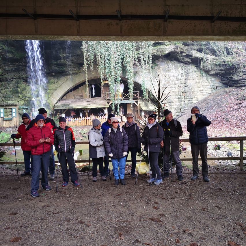 Eine Gruppe von Wanderern mit Trekkingstöcken steht unter einer Brücke, mit einem Wasserfall und einer Felsformation dahinter. Sie sind warm gekleidet für das kalte Wetter.