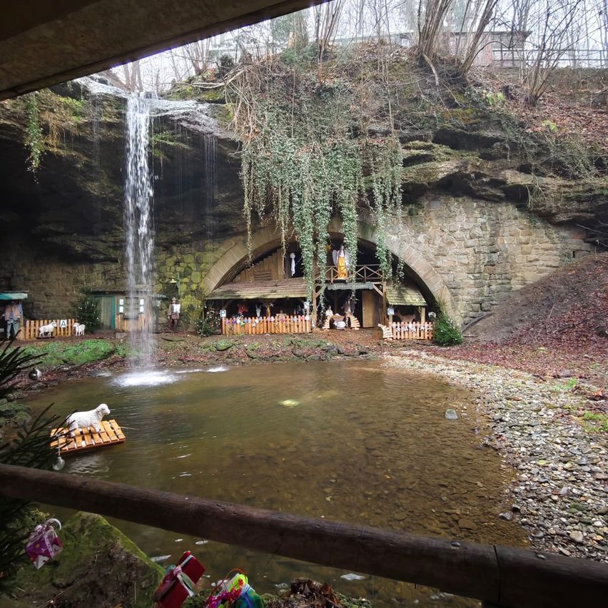 Ein Wasserfall stürzt von einer Klippe in einen Teich mit einem Holzfloß. Unter dem Wasserfall befindet sich ein steinerner Bogen, vor dem eine Krippe aufgestellt ist. Geschenke sind um den Teich herum platziert.