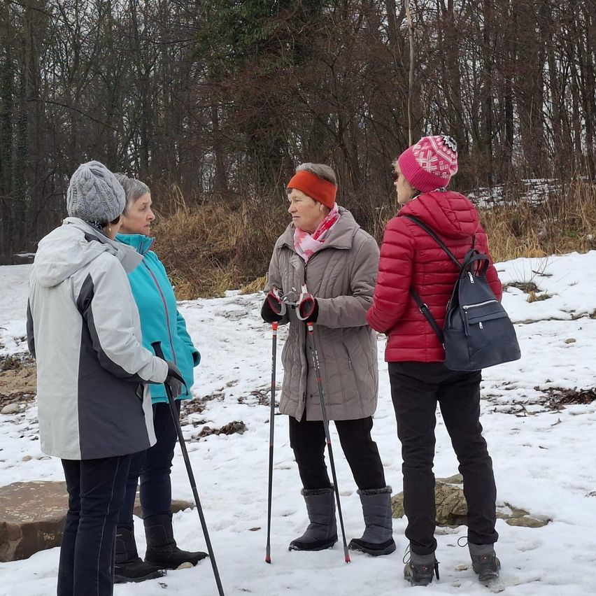 Vier Frauen in Winterkleidung stehen in einem verschneiten Feld und unterhalten sich. Zwei haben Skistöcke, eine hat eine Tasche. Dahinter säumen kahle Bäume einen Grasrand.