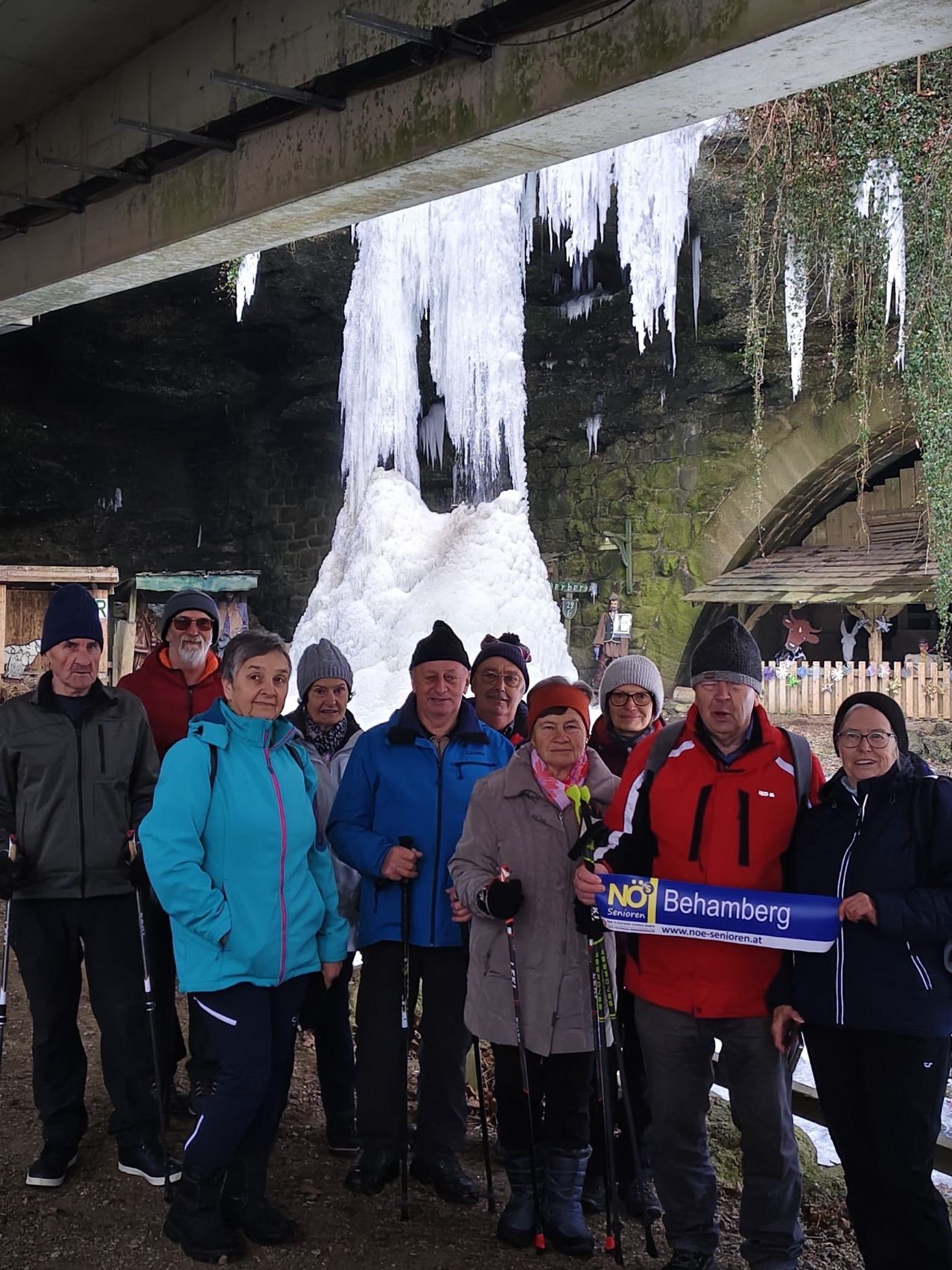 Eine Gruppe von Menschen in Winterkleidung posiert vor einem großen Eiszapfen. Im Hintergrund befinden sich eine Brücke und ein schneebedeckter Wasserfall.