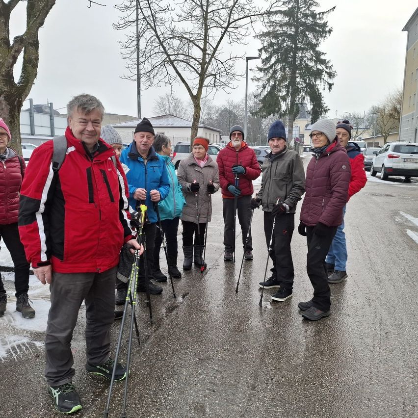 Eine Gruppe von Menschen in Winterkleidung steht auf einer nassen Straße, lächelt und hält Wanderstöcke in der Hand. Bäume und geparkte Autos säumen die Straße.
