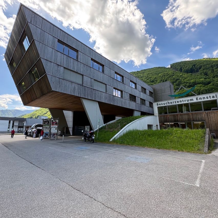 The Ennstal Visitor Center features a modern, angular building design. Several people are walking around outside, with a parked motorcycle nearby. Mountains are visible in the background.