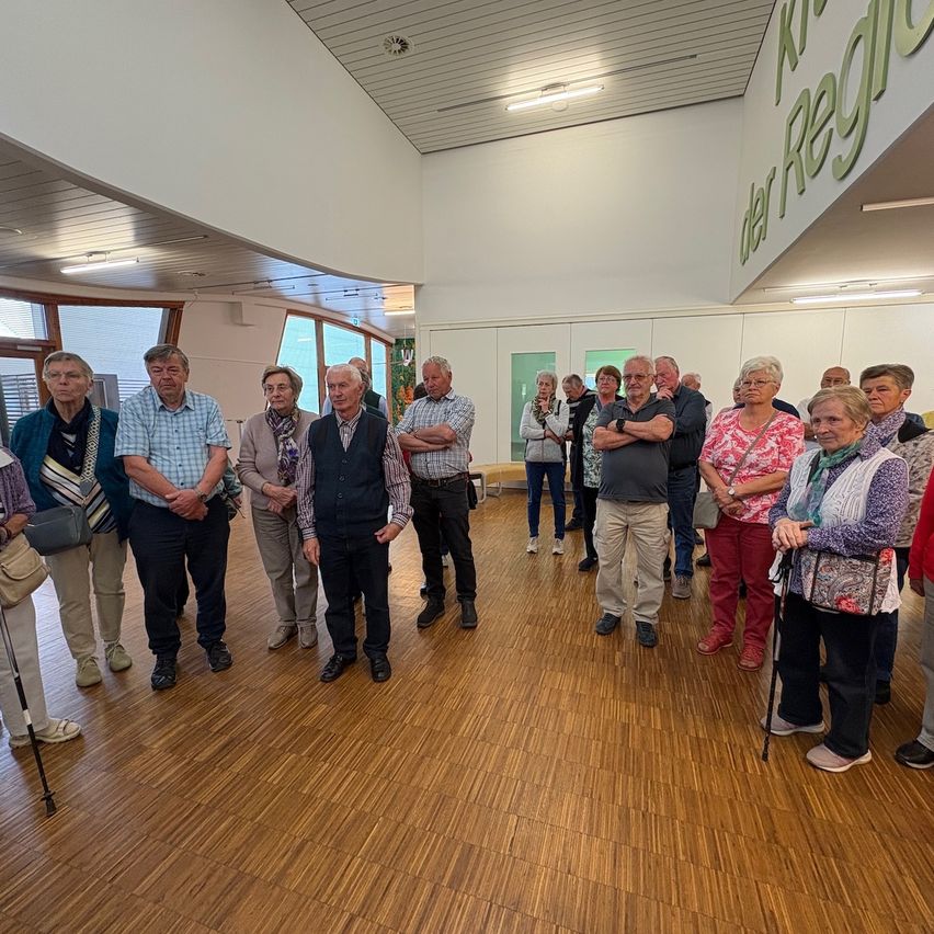 A group of older adults stands in a room with wooden floors. They are facing a sign that reads 'der Regi'.