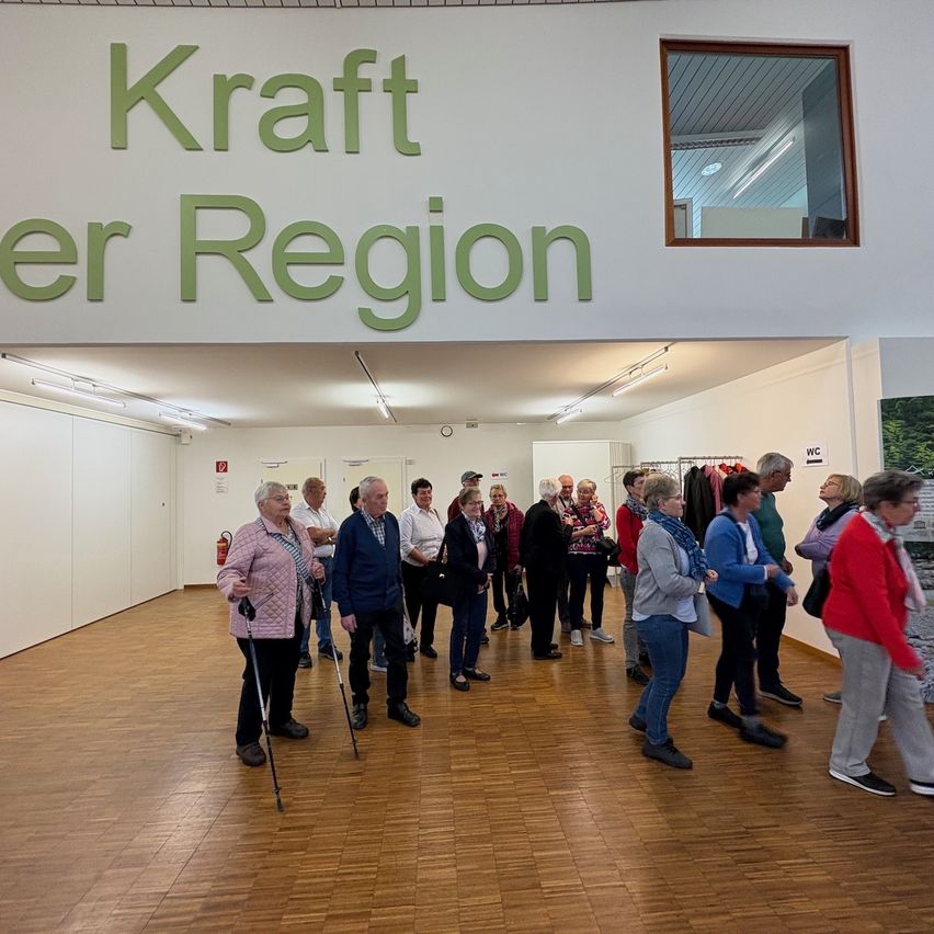 A group of people with walking sticks stand in an indoor area with wooden floors. The sign above reads 'Kraft er Region'.