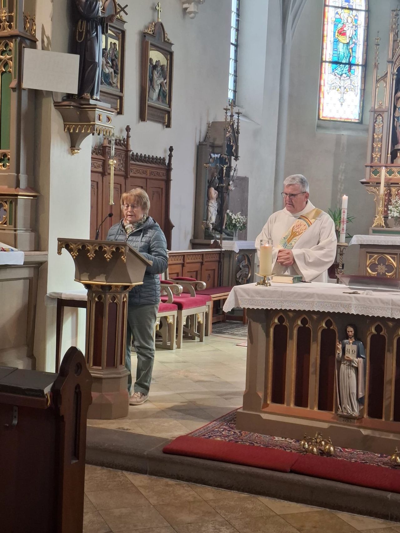 A priest stands by the altar with a candle while a woman reads from a podium in a church.