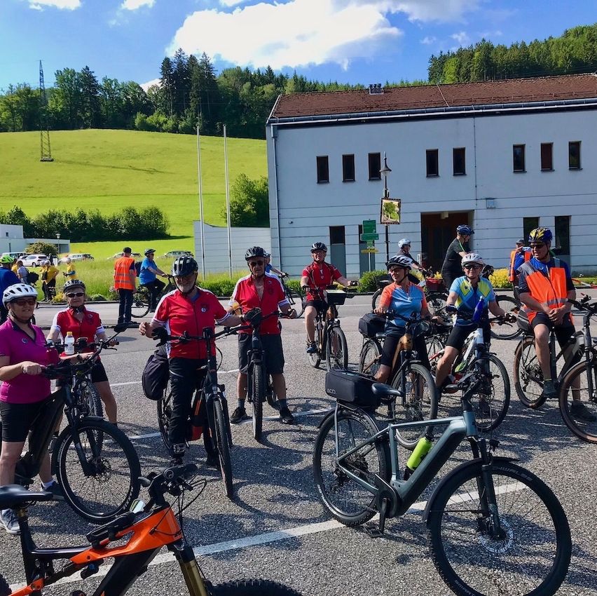 Eine Gruppe von Radfahrern mit Helmen und Fahrradausrüstung steht auf einem Parkplatz mit Fahrrädern, einem Gebäude und einem Feld im Hintergrund.