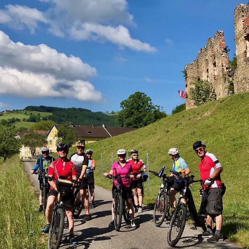 Eine Gruppe von Radfahrern mit Helmen fährt auf einer malerischen Route mit einer Burg im Hintergrund.
