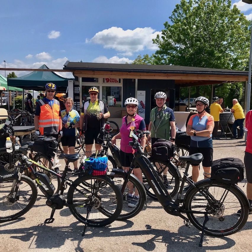 Eine Gruppe von Radfahrern posiert für ein Foto vor einem Gebäude, mit grünen Zelten und Bäumen im Hintergrund.