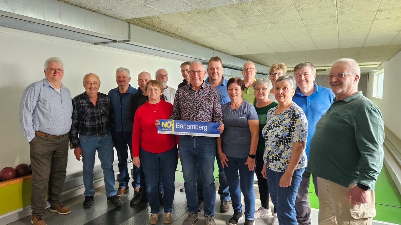 Eine Gruppe von Menschen steht in einem Raum und hält ein blaues Banner mit den Texten 'NEIN' und 'Behamberg'. Der Mann in der Mitte trägt eine Brille und ein Blumenhemd. Die anderen sind lässig gekleidet. Sie scheinen für ein Foto zu posieren.