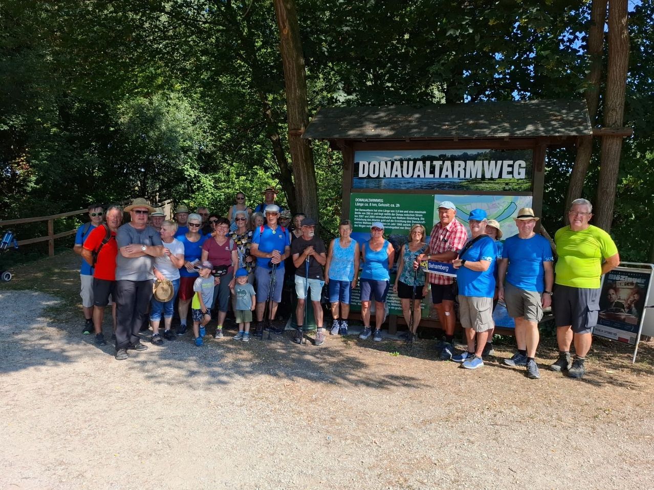 Eine Gruppe von Menschen in lässiger Sommerkleidung posiert für ein Foto vor einem Schild, das Donaualterarmweg steht. Das Schild ist an einer hölzernen Struktur mit Dach befestigt.