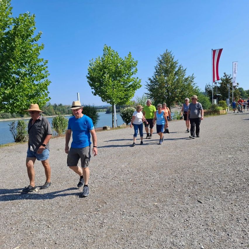 Eine Gruppe von Menschen geht auf einem Schotterweg in der Nähe eines Flusses, mit Bäumen und Fahnen im Hintergrund.